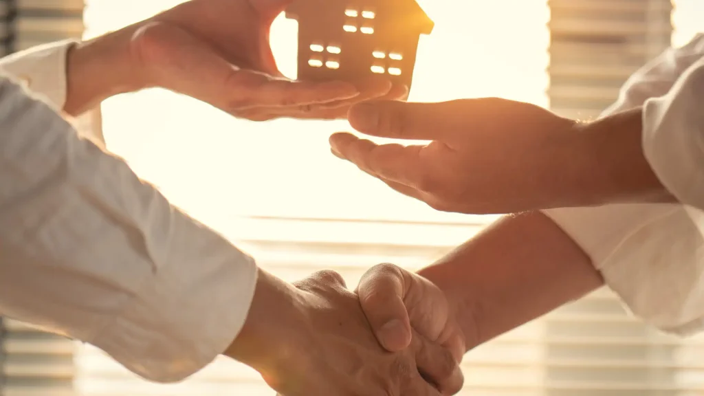 Handshake with person handing over small house model in warm lighting.